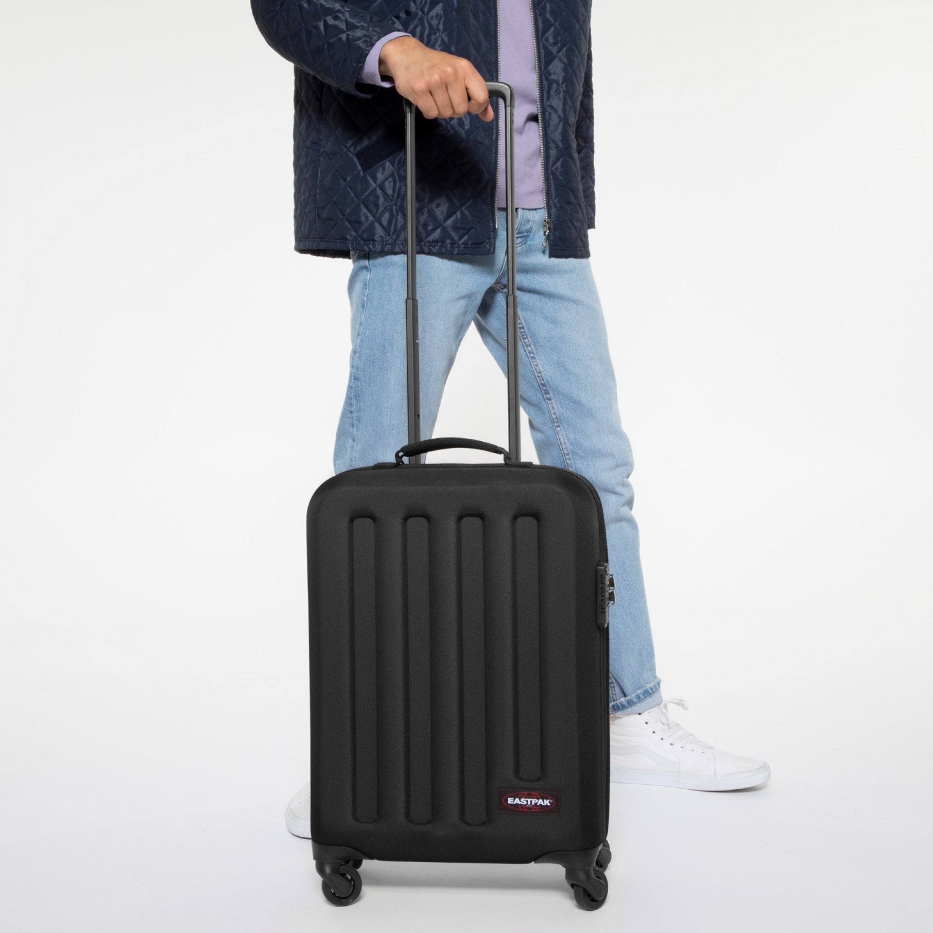 Person in jeans and a quilted jacket with a black Eastpak carry-on suitcase on four wheels